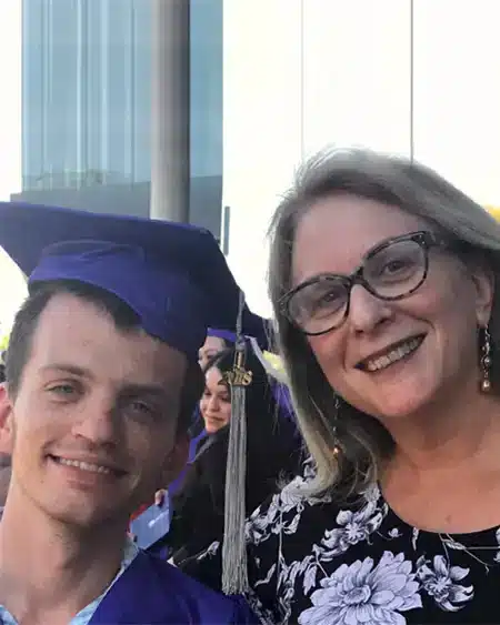 A young man in a graduation cap and gown stands next to his mother