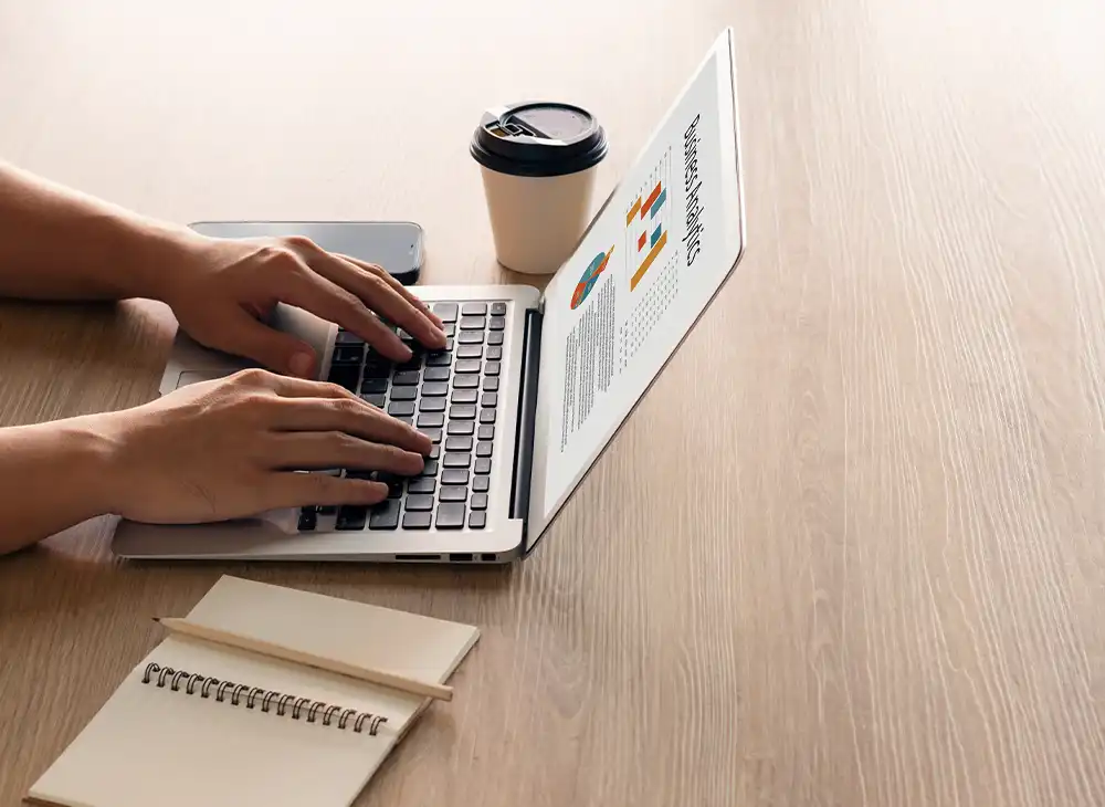 Hands typing on a laptop with graphs on the screen, a notebook, smartphone, and a coffee cup on the wooden table.